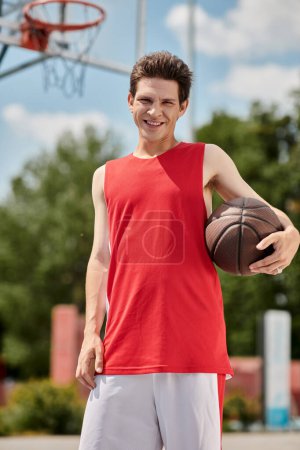 A young man in a red shirt skillfully holds a basketball on a sunny summer day outdoors.