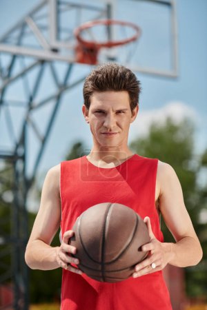Foto de Un joven con una camisa roja gotea hábilmente una pelota de baloncesto afuera en un día soleado de verano.. - Imagen libre de derechos