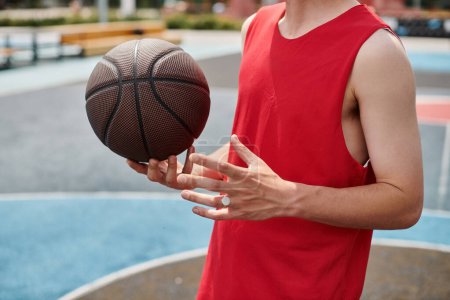 A young man in a vibrant red shirt happily holds a basketball outdoors on a sunny summer day.