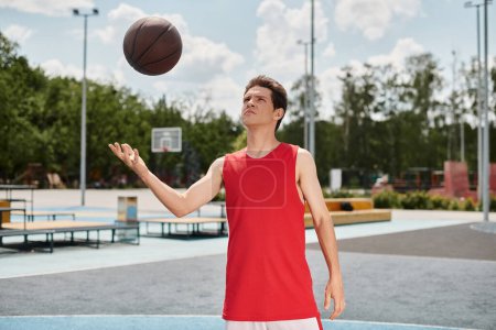 Młody koszykarz w czerwonej koszuli jest w połowie rzutu podczas gry w kosza na świeżym powietrzu w słoneczny letni dzień.