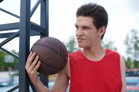 Un hombre con una camisa roja ardiente gotea hábilmente una pelota de baloncesto al aire libre en un día soleado de verano.