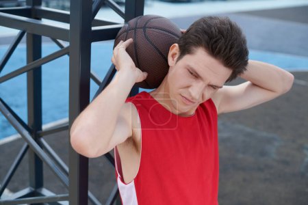 Un jeune homme en chemise rouge tient habilement un ballon de basket tout en se préparant à jouer à l'extérieur par une journée ensoleillée d'été.