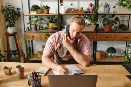 Un hombre se sienta en un escritorio hablando en un teléfono celular mientras trabaja en una tienda de plantas.