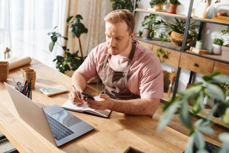 Un hombre trabajando en un portátil y un cuaderno en una mesa en un ambiente acogedor y lleno de plantas.