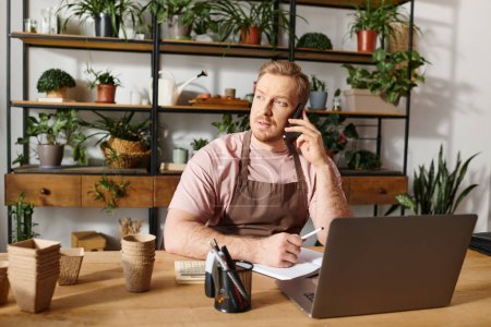 Un hombre se sienta en una mesa en una tienda de plantas, hablando por un teléfono celular mientras maneja su pequeño negocio.