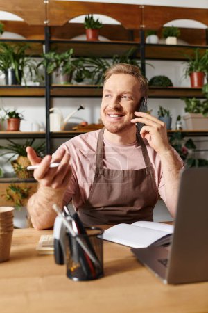 Un hombre sentado en una mesa en una tienda de plantas, hablando por un teléfono celular mientras está rodeado de vegetación.