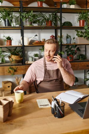 Un hombre se sienta en un escritorio en una tienda de plantas, hablando por un teléfono celular, mostrando determinación hacia el éxito de su pequeña empresa.