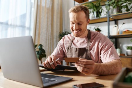 Un hombre sentado en una computadora portátil, sosteniendo una tarjeta de crédito, probablemente comprando suministros para su pequeño negocio de tiendas de plantas.