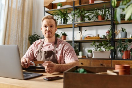 Foto de Un hombre está absorto en su portátil, rodeado de plantas en un entorno de pequeña empresa. - Imagen libre de derechos