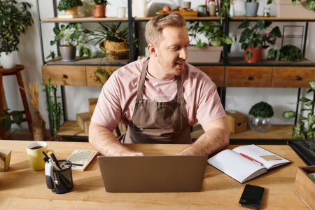 Foto de Un hombre centrado en su portátil en un escritorio, rodeado de plantas en un entorno de tienda de plantas. - Imagen libre de derechos