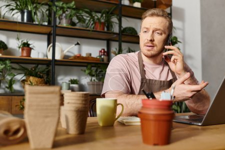 Foto de Un hombre se sienta en una mesa en una tienda de plantas, ocupado en una llamada telefónica. El entorno emana un ambiente de propietarios de pequeñas empresas. - Imagen libre de derechos