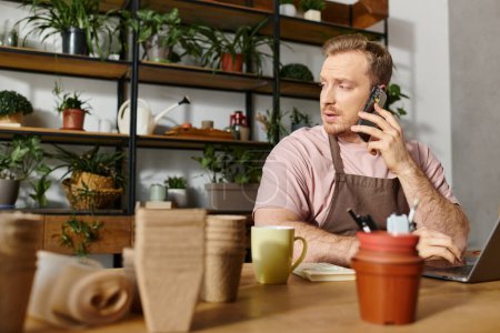 Ein Mann sitzt an einem Tisch in einem Pflanzenladen, vertieft in Gespräche auf seinem Handy, verkörpert einen Kleinunternehmer in Aktion.