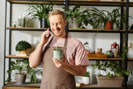 A man in an apron multitasks by holding a plant pot and talking on a cell phone in a botanical shop.