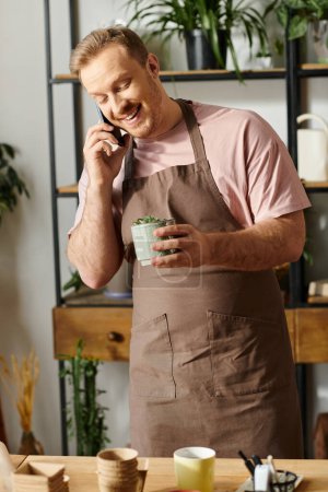 A handsome man in an apron multitasking as he converses on a cell phone in a plant shop setting.