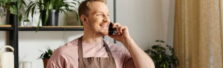Un hombre con estilo en un delantal conversa en un teléfono celular, administrando su tienda de plantas con un enfoque en el servicio al cliente.