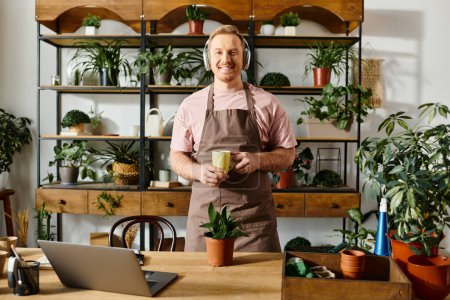 A handsome man in an apron holding a cup in front of a laptop in a plant shop setting.
