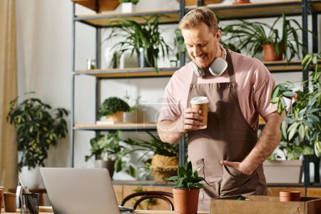 A man in a plant shop stands with a laptop, holding a cup of coffee as he begins his work day.