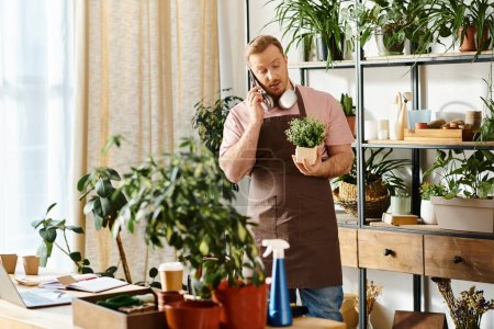 A man in an apron talks on a cell phone while holding a potted plant in a plant shop.