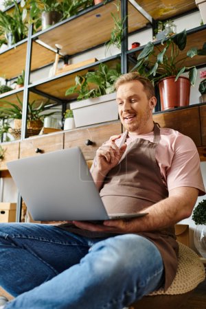 Foto de Un hombre con una computadora portátil se sienta en el suelo de una tienda de plantas, inmerso en su trabajo y rodeado de vegetación. - Imagen libre de derechos