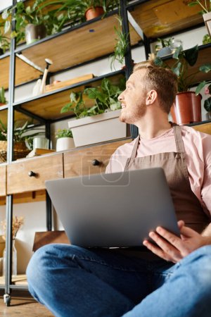 Un homme assis sur le sol, profondément concentré sur son ordinateur portable, entouré de plantes vertes luxuriantes dans un magasin de plantes.