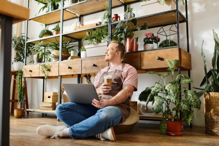 A stylish man sits on the floor, focused on his laptop at his small plant shop. He is busy managing his florist business.