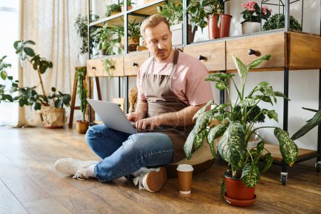 Foto de Un hombre en una tienda de plantas se sienta en el suelo trabajando intensamente en su computadora portátil, encarnando la dedicación a su pequeña empresa. - Imagen libre de derechos