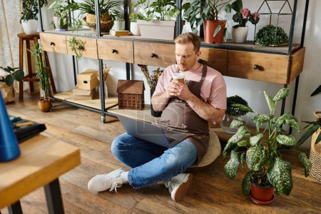 A man with a business shirt sits on floor, engrossed in laptop, surrounded by potted plants in a vibrant plant shop.