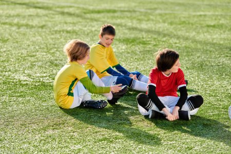 Téléchargez les photos : Un groupe diversifié de jeunes enfants sont joyeusement assis sur un terrain de soccer vert dynamique, coller et partager des rires ensemble dans la lumière du soleil doré de la fin de l'après-midi. - en image libre de droit