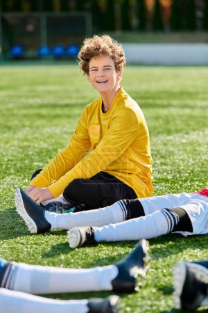 Téléchargez les photos : Un jeune garçon assis sur l'herbe avec les jambes croisées, en contemplation profonde sous le ciel bleu clair. - en image libre de droit