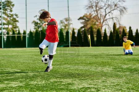 Téléchargez les photos : Un jeune garçon dynamique donne un coup de pied énergique à un ballon de football sur un terrain vert sous le soleil éclatant, montrant sa passion pour le sport et des compétences prometteuses. - en image libre de droit