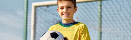 Téléchargez les photos : Un jeune garçon se tient fièrement debout, tenant un ballon de football devant un but. Avec détermination à ses yeux, il rêve un jour d'être un joueur étoile sur le terrain. - en image libre de droit