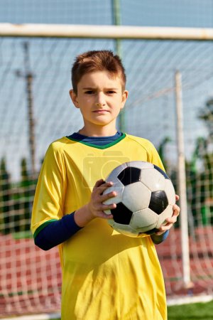 Ein kleiner Junge steht auf einer lebhaften grünen Wiese und hält einen Fußballball mit einem entschlossenen Blick im Gesicht..