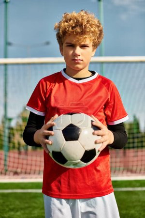 Ein kleiner Junge steht selbstbewusst auf einem riesigen Fußballfeld und wiegt einen Fußballball dicht an seiner Brust. Das leuchtend grüne Gras erstreckt sich um ihn herum, unter einem klaren blauen Himmel. Seine Augen leuchten vor Entschlossenheit und Aufregung, als er sich das Spiel vor Augen führt.