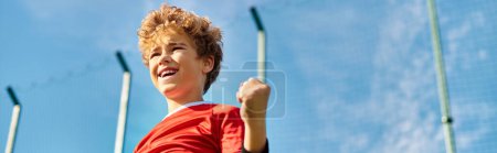 A boy in a vibrant red shirt confidently holds a baseball bat with a determined expression. He stands ready, showcasing his passion for the sport and readiness to swing.