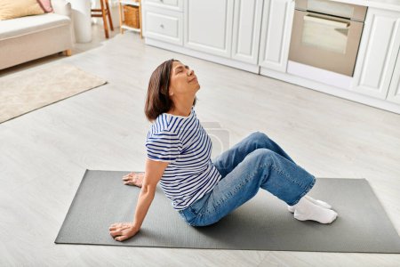 Reife Frau in gemütlicher Homewear praktiziert Yoga auf einer Matte in einem sonnigen Wohnzimmer.
