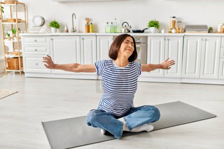Mature woman in cozy homewear sitting on a yoga mat, meditating in a sunlit kitchen.