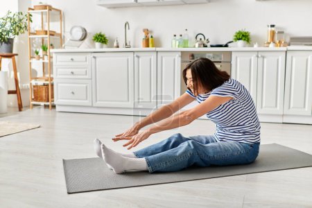 Eine reife schöne Frau in gemütlicher Hausbekleidung praktiziert Yoga auf einer Matte in ihrer Küche.