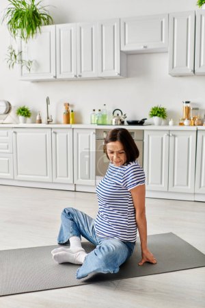 A mature, beautiful woman in cozy homewear sits on a yoga mat in the kitchen.