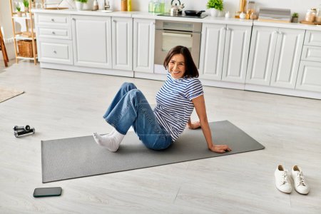 Femme mûre en vêtements de maison pratique le yoga sur un tapis dans une cuisine confortable.