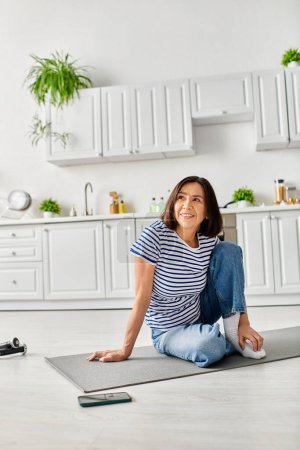 Téléchargez les photos : Une femme mature et belle dans des vêtements confortables pratiquant le yoga sur un tapis dans sa cuisine. - en image libre de droit