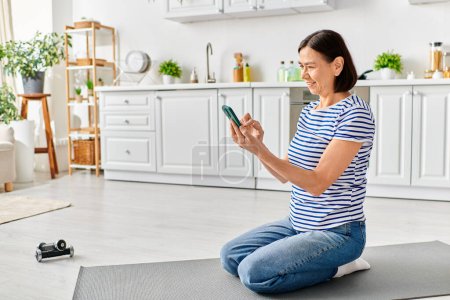 Reife Frau in gemütlicher Kleidung sitzt auf Yogamatte und konzentriert sich aufs Telefon.
