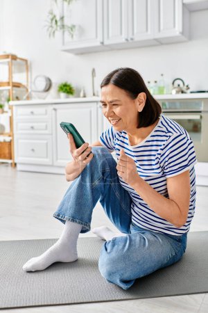 Eine reife Frau in kuscheliger Homewear sitzt auf dem Fußboden, in ihr Handy vertieft.