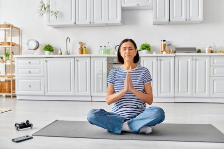 Téléchargez les photos : Une femme mature et belle dans des vêtements confortables pratiquant le yoga sur un tapis dans sa cuisine. - en image libre de droit