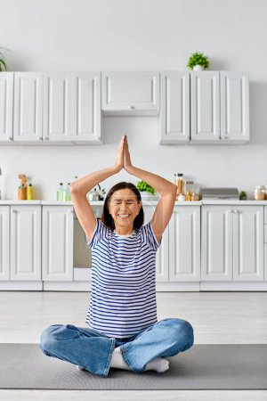 Téléchargez les photos : Femme mûre dans des vêtements confortables, assis sur un tapis de yoga avec les mains dans l'air. - en image libre de droit