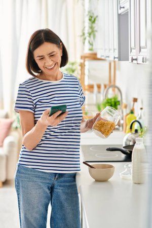 Mature woman in cozy homewear holding cell phone in kitchen.