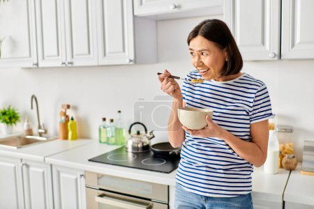 A mature woman in cozy homewear enjoys a bowl of cereal in her kitchen.