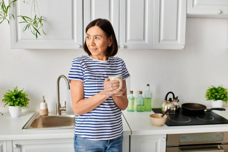 A mature woman in cozy homewear standing in a kitchen, holding a cup.