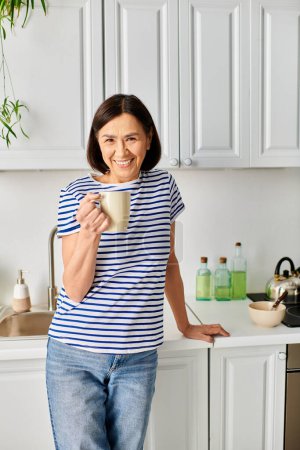 A woman in cozy homewear stands in a kitchen, holding a cup.
