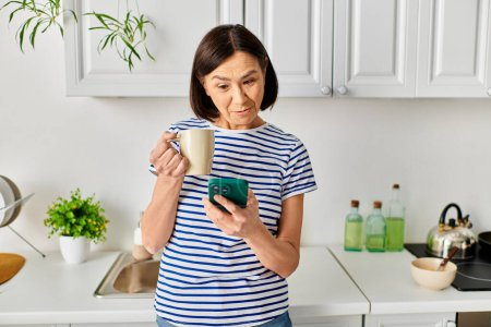 Una mujer en ropa de casa acogedora de pie en una cocina, sosteniendo una taza y un teléfono celular.