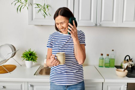 Una mujer madura en ropa de casa acogedora de pie en una cocina, charlando en un teléfono celular.